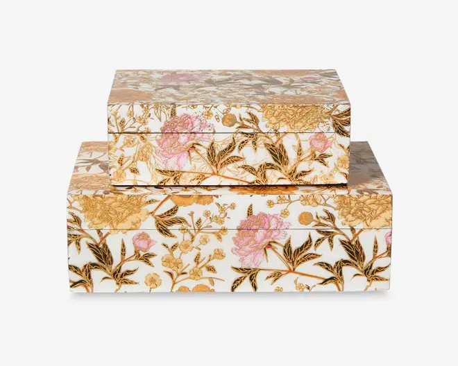 Sæt med to rektangulære opbevaringskasser, hver med en blank overflade og et detaljeret blomstermønster i guld, lyserød og grønne nuancer. Kasserne har et sømløst design, hvor blomstermotivet fortsætter rundt om alle sider, og blomsterne har fine blade og blomsterhoveder. Den større kasse fungerer som base, mens den mindre kasse er stablet ovenpå. Begge kasser har hængslet låg og er designet til dekorativ opbevaring.