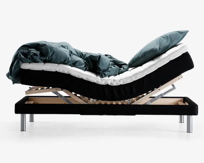 Senge set fra siden med en justerbar bund, hvor både hoved- og fodende er hævet. Sengen har en sort ramme med metalben, en hvid madras og mørkegrønt sengetøj. Den lamelbaserede træramme under madrassen er synlig og viser sengens justerbare funktion.