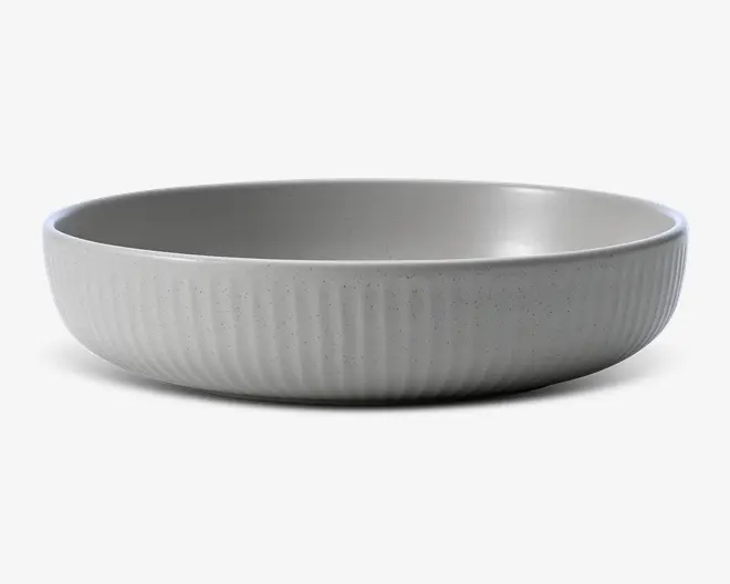 En lav, rund skål med mat, lysegrå overflade. Ydersiden har diskrete lodrette riller, der giver ekstra struktur, mens indersiden er glat. Skålen har et minimalistisk og moderne design med en let plettet overflade, der giver et håndlavet udtryk.
