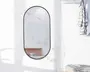 Et lodret, ovalt spejl med en tynd sort metalramme. Spejlet er monteret på en hvid væg og reflekterer en overflade med marmormønster samt en træhylde med et par stablede bøger. Designet er minimalistisk og moderne, velegnet til nutidige indretninger.