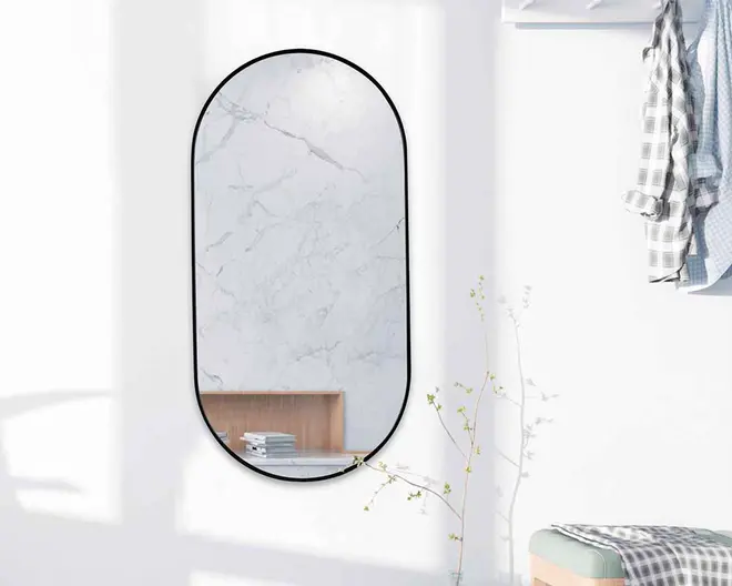 Et lodret, ovalt spejl med en tynd sort metalramme. Spejlet er monteret på en hvid væg og reflekterer en overflade med marmormønster samt en træhylde med et par stablede bøger. Designet er minimalistisk og moderne, velegnet til nutidige indretninger.