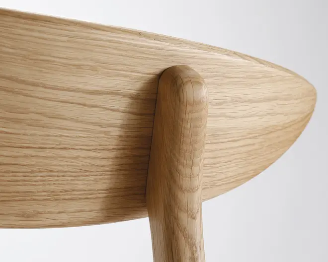 Nærbillede af ryglænet og støtten på en træstol, der fremhæver de glatte, afrundede kanter og den naturlige, lyse egetræsfinish. Træets åretegning er tydelig, hvilket understreger det fine håndværk og den sømløse samling mellem ryglæn og den lodrette støtte.