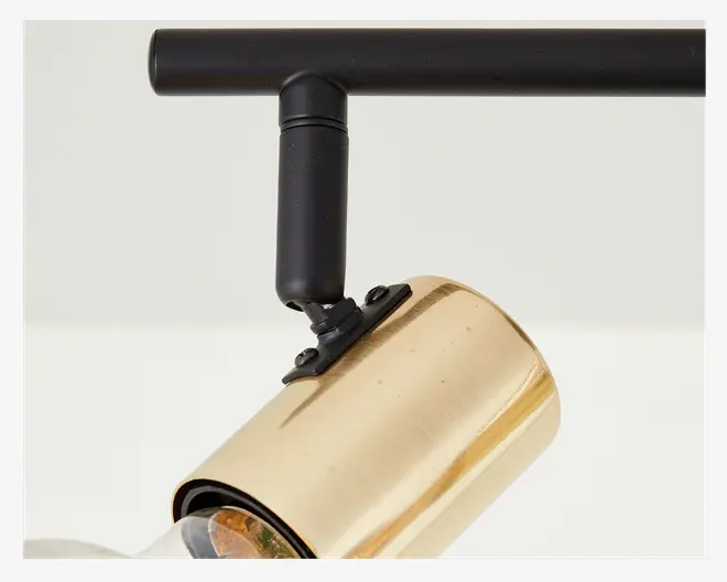 Nærbillede af en cylindrisk lampe med børstet guldfinish, monteret på en mat sort justerbar arm. Lampen har et drejeled, der gør det muligt at rette lyset, og der er en synlig pære inde i det gyldne lampehus.