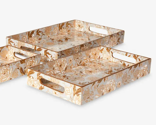 Sæt med tre rektangulære bakker i forskellige størrelser, hver med en blank overflade og et detaljeret guld- og hvidt blomstermønster. Bakkerne har høje kanter og indbyggede ovale udskæringer som håndtag i de korte ender for nem transport. Blomstermotivet dækker både inderside og yderside, hvilket giver et sammenhængende og elegant udtryk.