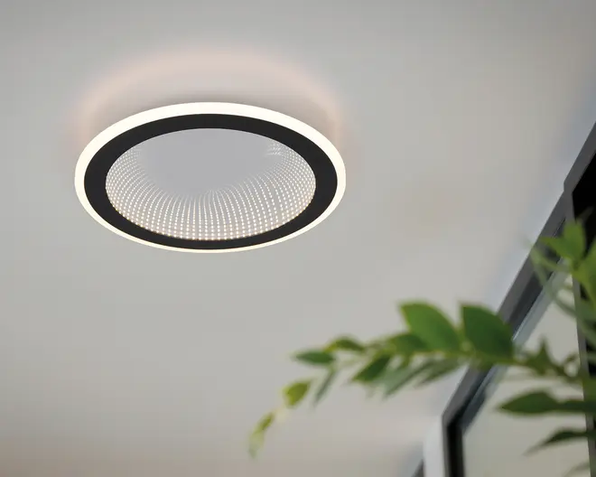 Moderne loftlampe med et cirkulært design, der har en sort inderring og en hvid yderring. Midten har et tekstureret, prikket mønster, som skaber en subtil optisk illusion og giver dybde og visuel interesse. Lampen udsender et blødt, diffust lys, der passer godt til moderne indretning.