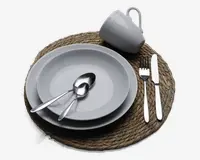 Et borddækning med en rund vævet dækkeserviet, en hvid keramisk middagstallerken og en mindre tallerken stablet ovenpå, to spiseskeer af rustfrit stål placeret på tallerkenerne, en hvid keramisk kop placeret over tallerkenerne samt et sæt bestik (gaffel og kniv) af rustfrit stål arrangeret til højre for tallerkenerne. Udtrykket er enkelt og moderne med neutrale farver og en naturlig tekstur fra dækkeservietten.