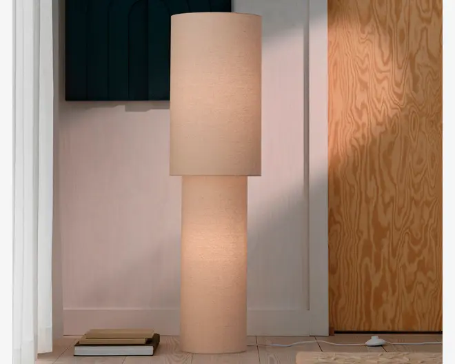 Høj cylindrisk gulvlampe med beige stofskærm og matchende beklædt fod, der afgiver et varmt, blødt lys.