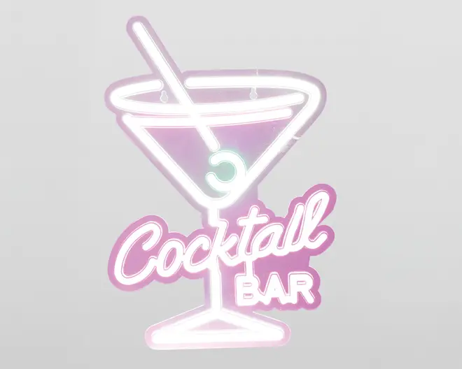 Neonskilt med et martiniglas, en rørepind og en oliven, omkranset af lyserøde og hvide lys. Teksten 'Cocktail Bar' står under glasset, hvor 'Cocktail' er skrevet med stor, kursiv skrift, og 'BAR' står med blokbogstaver. Designet har et retro og legende udtryk med en blød glød.
