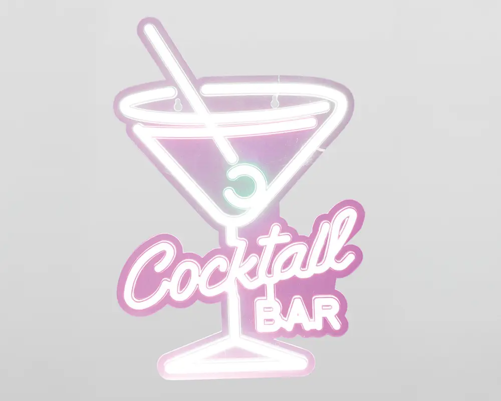 Neonskilt med et martiniglas, en rørepind og en oliven, omkranset af lyserøde og hvide lys. Teksten 'Cocktail Bar' står under glasset, hvor 'Cocktail' er skrevet med stor, kursiv skrift, og 'BAR' står med blokbogstaver. Designet har et retro og legende udtryk med en blød glød.