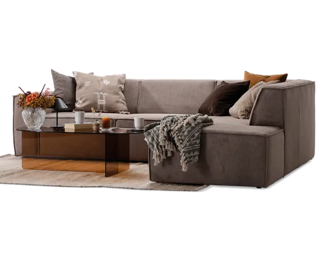 En moderne L-formet hjørnesofa i en neutral taupe farve med brede, lavprofilerede armlæn og bløde sædehynder. Sofaen er pyntet med forskellige dekorative puder i beige, brun og cremefarver samt et tekstureret, frynset tæppe, der er lagt over et hjørne. Foran sofaen står et stilfuldt, rektangulært sofabord i glas med bronzefarvet base, hvorpå der står en vase med friske blomster, stearinlys, bøger og kopper. Hele opstillingen står på et lysefarvet tæppe og skaber en hyggelig og indbydende stue.