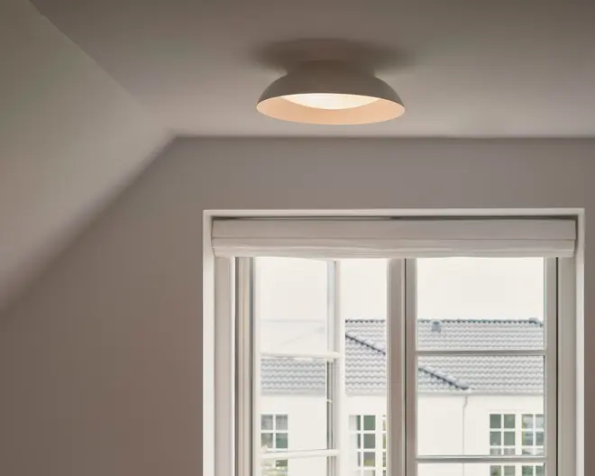 Mat grå, kuppelformet loftarmatur med varm, lysende diffuser, vist monteret tæt op ad et hvidt loft i et neutralt farvet rum.