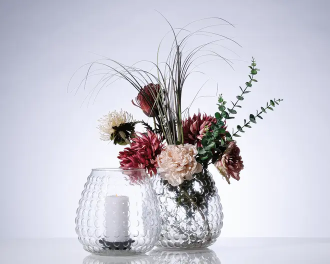 To klare glasvaser med en boblet tekstur. Den ene vase indeholder et hvidt bloklys, mens den anden har en buket kunstige blomster i bordeaux, creme og grønne nuancer samt lange dekorative græsstrå og blade. Vaserne har en rund form og et moderne, elegant udtryk.