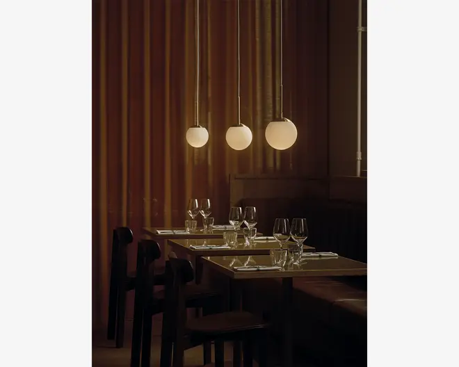 Tre pendellamper med matteret glas i kugleform hænger fra loftet og udsender et varmt, blødt lys. Lamperne er ophængt i slanke, metalliske stænger og er jævnt fordelt over et spiseområde. Bordene er dækket med vinglas, vandglas og pænt foldede servietter, hvilket skaber en elegant og indbydende stemning. Baggrunden består af gulv-til-loft gardiner i en afdæmpet brun farve, som bidrager til den hyggelige atmosfære.