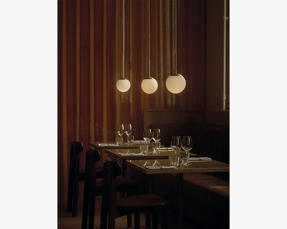 Tre pendellamper med matteret glas i kugleform hænger fra loftet og udsender et varmt, blødt lys. Lamperne er ophængt i slanke, metalliske stænger og er jævnt fordelt over et spiseområde. Bordene er dækket med vinglas, vandglas og pænt foldede servietter, hvilket skaber en elegant og indbydende stemning. Baggrunden består af gulv-til-loft gardiner i en afdæmpet brun farve, som bidrager til den hyggelige atmosfære.