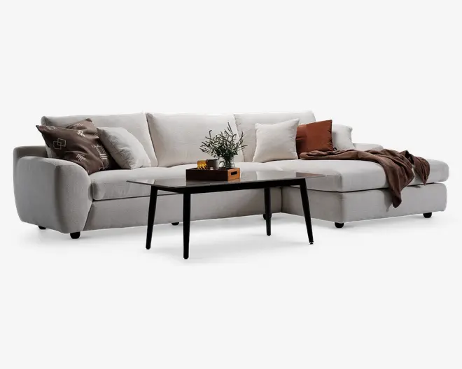 Moderne L-formet sofa betrukket med lysebeige stof, med brede armlæn og bløde ryghynder. Sofaen er pyntet med forskellige dekorative puder i brune, beige og rustfarver samt et blødt brunt tæppe lagt over den ene side. Foran sofaen står et elegant sort sofabord med en lille plante, en bog og en dekorativ æske.