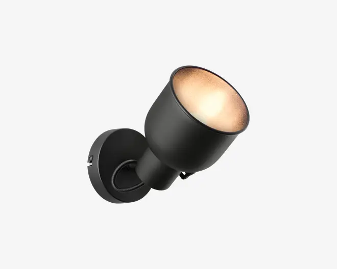 Et moderne vægmonteret spotlys med mat sort finish. Lampen har en rund base og en konisk skærm, der leder lyset fremad. Designet er minimalistisk med rene linjer og en glat overflade, velegnet til moderne indretning. Lyskilden er forsænket i skærmen, hvilket giver et fokuseret lys.