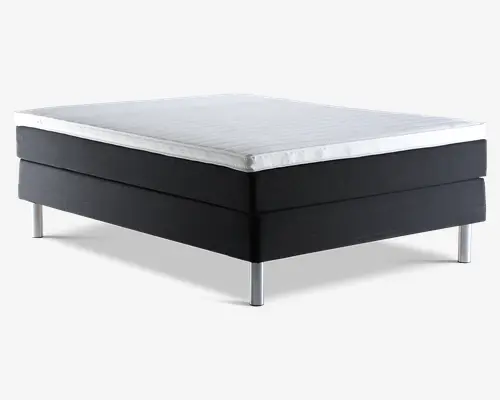 Rektangulær sengebund med sort polstret ramme og en hvid, quiltet topmadras. Sengen står på seks cylindriske sølvfarvede metalben, som giver solid støtte og et moderne udtryk. Topmadrassens kanter er pænt afsluttede, og det samlede design er minimalistisk og moderne.