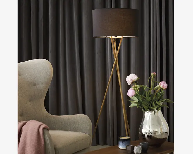 En moderne gulvlampe med en sort, trommeformet lampeskærm og tre slanke, guldfarvede ben arrangeret i et stativdesign. Lampen står ved siden af en lysegrå polstret lænestol med høj ryg og buede sider. Et blødt lyserødt tæppe er lagt over armlænet på stolen. Foran lampen står et træbord med en reflekterende sølvvase, der indeholder lyserøde pæoner og grønt løv, samt flere små dekorative genstande.