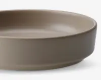 Et nærbillede af en rund, lav skål med en glat, mat overflade i en neutral beige farve. Skålen har lige, lidt tykke sider og en flad bund, hvilket giver den et moderne og minimalistisk udtryk.