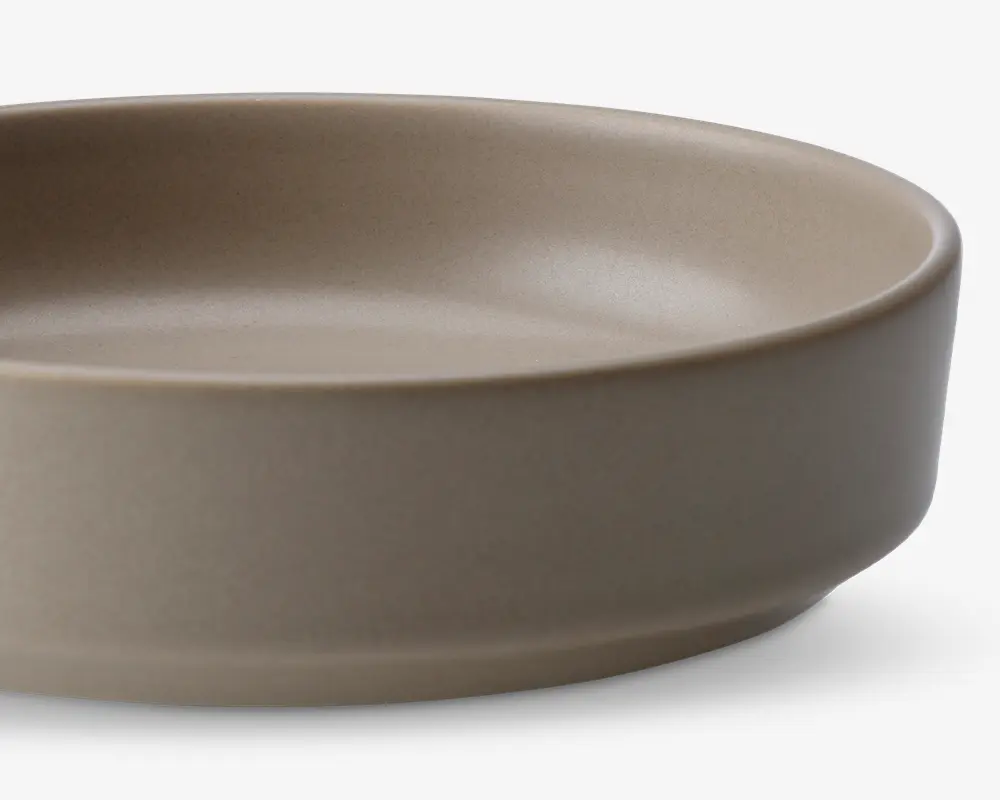 Et nærbillede af en rund, lav skål med en glat, mat overflade i en neutral beige farve. Skålen har lige, lidt tykke sider og en flad bund, hvilket giver den et moderne og minimalistisk udtryk.