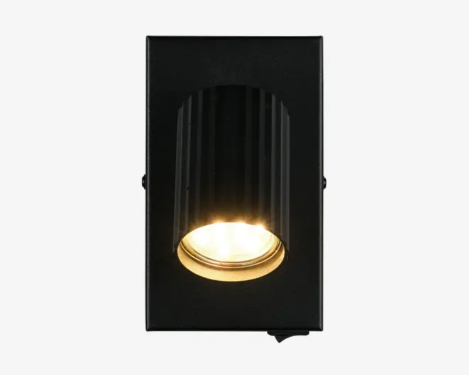 Rektangulær vægmonteret lampe med mat sort finish og en lodret rillet, cylindrisk lampeholder. Lyskilden er forsænket og udsender et varmt, fokuseret lys nedad. Lampens inderkant har en guldfarvet tone, som giver et elegant og kontrastfyldt udtryk. Egnet til moderne indretning, hvor lampen kombinerer dekorativt og funktionelt lys.