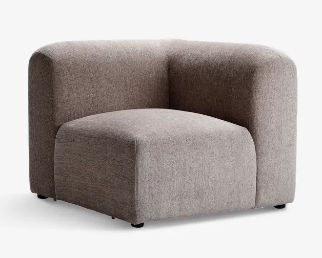 En moderne hjørnestol med minimalistisk design, betrukket med blødt, tekstureret lysebrunt stof. Stolen har en tyk, polstret siddehynde og afrundede ryg- og armlæn, der danner en glat, sammenhængende kurve. Basen er lav, og den samlede form er kantet, hvilket giver et moderne og hyggeligt udtryk.