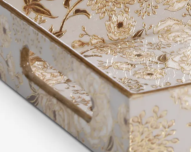 Rektangulær bakke med blank hvid overflade, dekoreret med indviklede, hævede guldfarvede blomster-mønstre. Bakken har udskårne håndtag i siderne for nem bæring, og gulddetaljerne fortsætter langs kanter og håndtag, hvilket giver et elegant og luksuriøst udtryk.