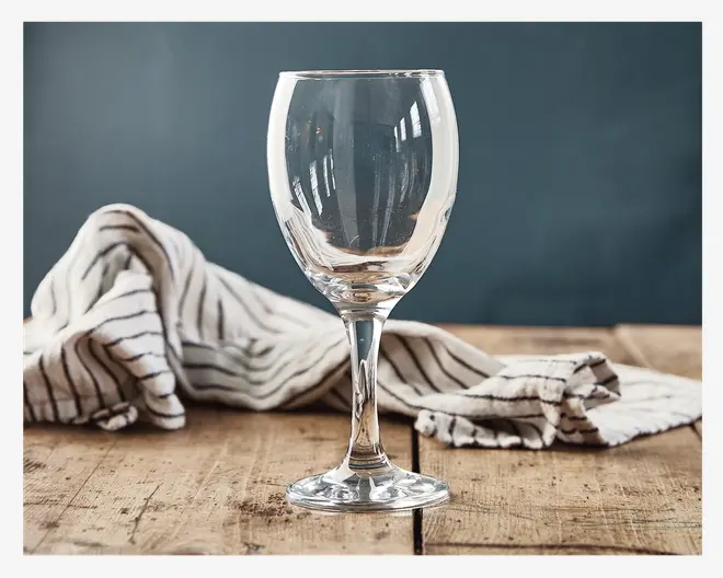 Klar glas med rundet skål, slank stilk og flad fod. Glasset er gennemsigtigt og har en glat overflade, velegnet til servering af vin eller lignende drikkevarer. Det står på et rustikt træbord med stribede klude i baggrunden.