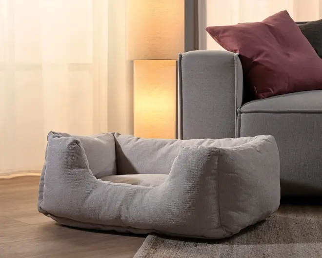 Blød lysegrå rektangulær kæledyrsseng med polstrede sider, lav åbning foran og en forsænket liggeflade, vist på et trægulv ved siden af en sofa.
