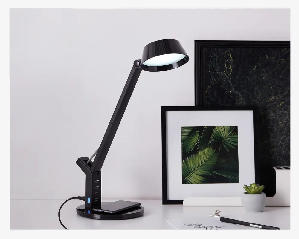 Elegant, moderne sort skrivebordslampe med justerbar arm og rundt lampehoved. Lampen har et touch-kontrolpanel i basen, som også indeholder USB-opladningsporte. Lampen er tændt og har en solid, vægtet base for stabilitet. Designet er minimalistisk og passer til moderne arbejdsområder.