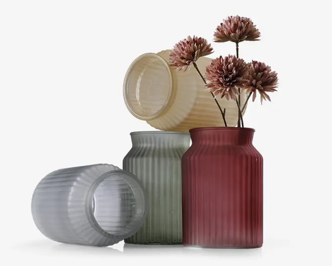 Fire rillede glasbeholdere i forskellige farver – mat hvid, lysegul, lysegrøn og mørkerød. Alle har en cylindrisk form med lodrette riller, der giver tekstur. Den mørkerøde beholder står oprejst og indeholder flere kunstige lyserøde blomster, mens de andre er tomme og ligger ned eller står op. Glasset har en mat finish, hvilket giver et blødt og elegant udtryk.