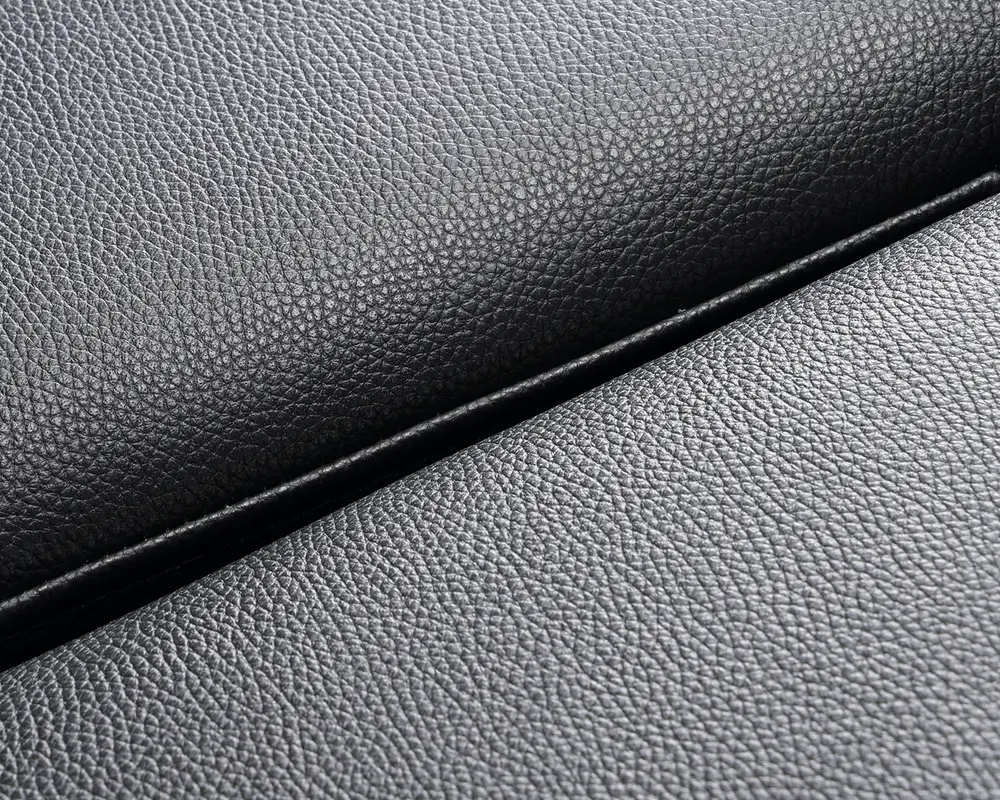 Nærbillede af et sort materiale med en fin, småkornet tekstur og mat finish. Overfladen ser smidig og fleksibel ud med en let glans, der fremhæver kornmønsteret. Billedet viser en foldet kant, hvilket indikerer materialets tykkelse og bøjelighed.