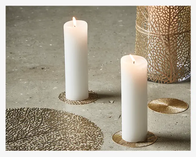 To høje, hvide bloklys er tændt og står oprejst på en glat, grå overflade. Hvert lys står på en dekorativ, rund bordskåner i guldfarvet metal med et tekstureret, indviklet mønster. I baggrunden ses en guldfarvet lanterne med et bladmønster i udskæringer, og i forgrunden er en matchende guldfarvet dækkeserviet med lignende detaljer delvist synlig.