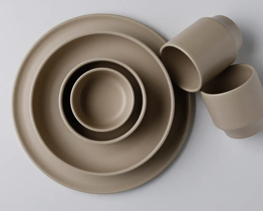 Et sæt mat beige service bestående af to runde tallerkener i forskellige størrelser, en lille skål og to kopper. Designet er minimalistisk med glatte, rene linjer og ensartet farve. Delene kan stables og har et moderne, diskret udtryk.