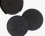 Sæt af runde, sorte coasters i kunstlæder med synlige syninger langs kanten. Hver coaster har præget, humoristisk tekst: én med teksten 'WINE IS CHEAPER THAN THERAPY' og en anden med 'WHISKEY GIVES YOU BALLS.' Sættet leveres med en matchende holder til opbevaring.