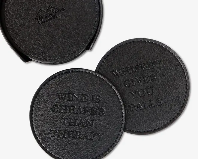 Sæt af runde, sorte coasters i kunstlæder med synlige syninger langs kanten. Hver coaster har præget, humoristisk tekst: én med teksten 'WINE IS CHEAPER THAN THERAPY' og en anden med 'WHISKEY GIVES YOU BALLS.' Sættet leveres med en matchende holder til opbevaring.
