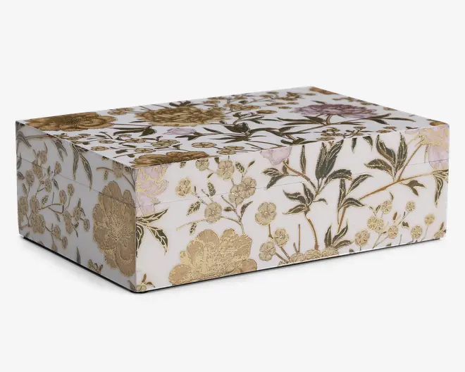 Rektangulær æske med hængslet låg, dekoreret med et hvidt baggrund og et detaljeret blomstermønster i guld, grøn og diskrete lavendelfarver. Blomsterne og bladene er fint udført, og nogle elementer har en metallisk guldfinish, der giver et elegant og luksuriøst udtryk. Æsken har skarpe kanter og en glat, blank overflade.