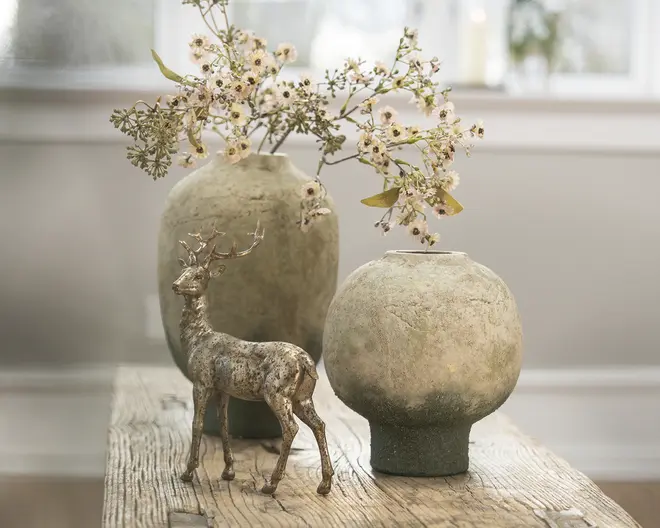 To runde keramikvaser med ru overflade i dæmpede beige- og olivenfarver står på en slidt træflade, den ene høj og den anden lav, med lyse, fine blomster i den højeste vase og en lille sølvfarvet hjortefigur foran.
