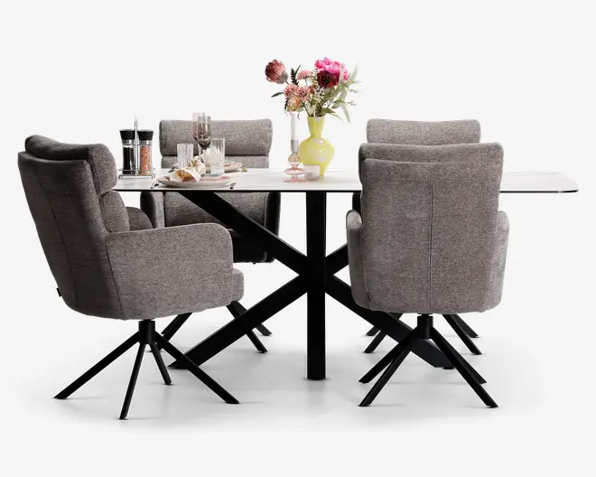Et moderne spisesæt bestående af et rektangulært bord med lys bordplade og sorte metalben i krydsdesign. Sættet inkluderer seks polstrede stole betrukket med blødt gråt stof, alle med armlæn og sorte metalben med drejefunktion. Bordet er pyntet med en gul vase med en buket lyserøde og røde blomster, glas, tallerkener og en stempelkande, hvilket skaber en indbydende og elegant spiseatmosfære.