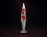 En høj, raketformet lampe med en metallisk sølvfarvet fod og top. Den gennemsigtige midtersektion indeholder røde voksbobler, der flyder i en klar væske og skaber en dynamisk, bevægelig effekt, når lampen er tændt. Lampen får strøm via en ledning, der er synlig ved foden.