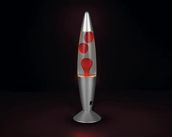 En høj, raketformet lampe med en metallisk sølvfarvet fod og top. Den gennemsigtige midtersektion indeholder røde voksbobler, der flyder i en klar væske og skaber en dynamisk, bevægelig effekt, når lampen er tændt. Lampen får strøm via en ledning, der er synlig ved foden.