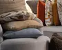 Stablede pyntepuder i neutrale og varme farver, herunder beige striber, tekstureret taupe, blød blågrå, rustorange og mønstrede cremefarvede puder arrangeret på en sofa.