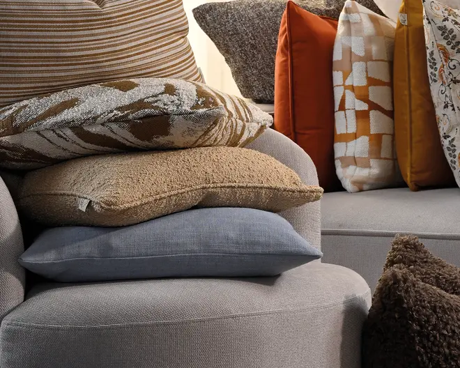 Stablede pyntepuder i neutrale og varme farver, herunder beige striber, tekstureret taupe, blød blågrå, rustorange og mønstrede cremefarvede puder arrangeret på en sofa.