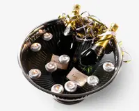 En stor, rund metalskål fyldt med vand, indeholdende to flasker mousserende vin med gyldne folie-toppe og dekorative guld- og sølvbånd. Flere små fyrfadslys flyder på vandet omkring flaskerne, hvilket giver en festlig og elegant præsentation.