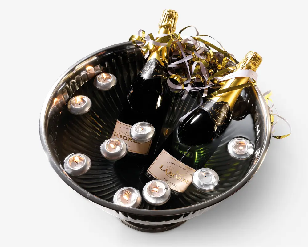En stor, rund metalskål fyldt med vand, indeholdende to flasker mousserende vin med gyldne folie-toppe og dekorative guld- og sølvbånd. Flere små fyrfadslys flyder på vandet omkring flaskerne, hvilket giver en festlig og elegant præsentation.