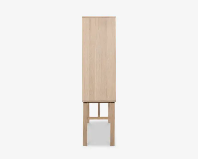 Sidevisning af et højt, smalt skab lavet af lyst træ. Skabet står på fire lige ben med et rektangulært åbent rum under hovedopbevaringsdelen, hvilket giver et løftet og let udtryk. Træets årer er synlige, og designet er minimalistisk med rene linjer og uden synlige greb eller dekorative elementer.