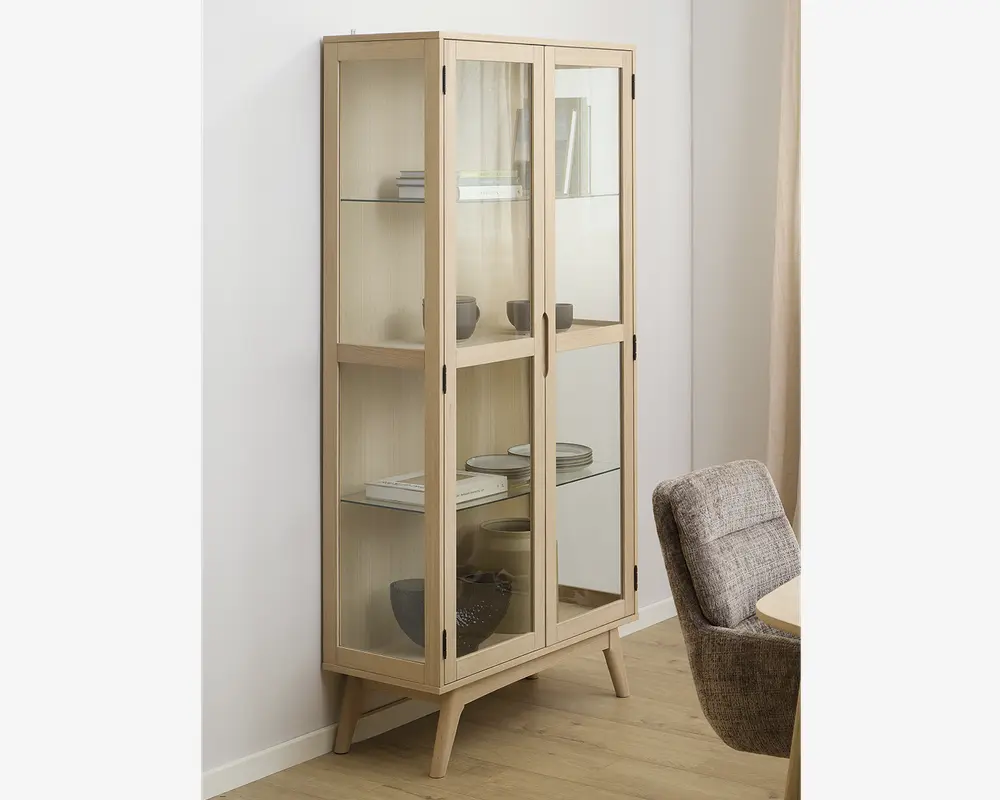 Høj vitrineskab med lys træramme og glaslåger, udstyret med tre glashylder til opbevaring. Skabet står på fire skråtstillede ben, hvilket giver et moderne, retro-inspireret udtryk. Indretningen er rummelig og gør det nemt at se de placerede genstande, såsom bøger, skåle og dekorative objekter.