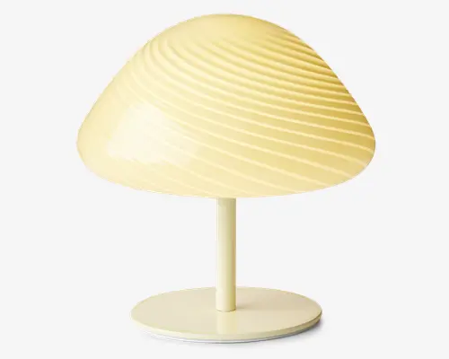 Bordlampe med en kuppelformet skærm i en blød cremefarve. Skærmen har en markant spiralformet rillet struktur, som giver lampen et moderne og stilfuldt udtryk. Lampen har en enkel cylindrisk stander og en rund, flad fod, begge i samme farve som skærmen for et ensartet udseende.