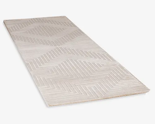 Rektangulært tæppe i en lys beige farve med et moderne geometrisk mønster. Designet består af chevron- og zigzag-linjer, der giver en tekstureret, tredimensionel effekt. Tæppet har en lav luv og et enkelt, minimalistisk udtryk, der passer til moderne indretning.