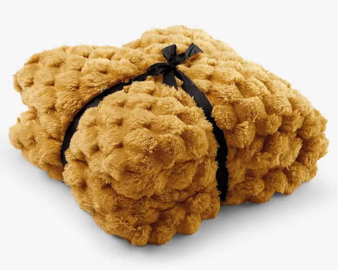Gyldenbrun, blød plaid i imitation pels med kraftig, luftig struktur og sort bånd, der holder den sammenfoldede pakke samlet.